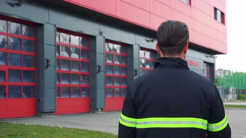 Ein Feuerwehrmann (Der Rücken zur Kamera) schaut auf eine Feuerwache und setzt einen Helm auf