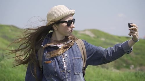 A Woman with Long Hair in a Hat and Sunglasses is Filming Herself in Nature