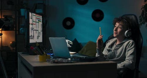 A Boy Sits in Room in the Evening in Front of Computer Screen and Laptop Working on a School Project