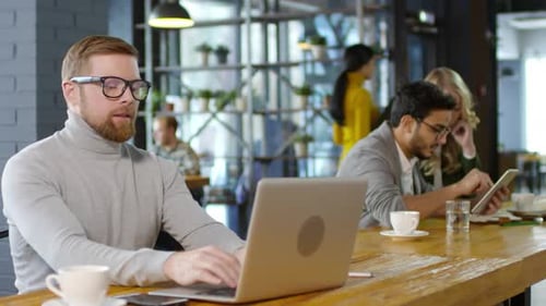 Fröhlicher kaukasischer Mann tippt auf Laptop und posiert vor der Kamera im Café