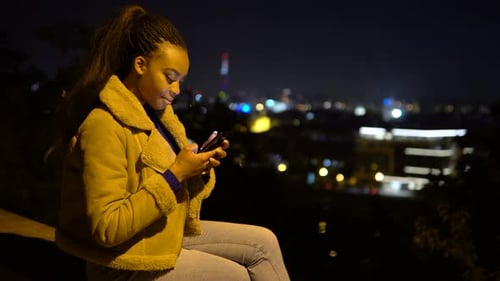 A Young Black Woman Takes Selfies with a Smartphone As She Sits in an Urban Area at Night
