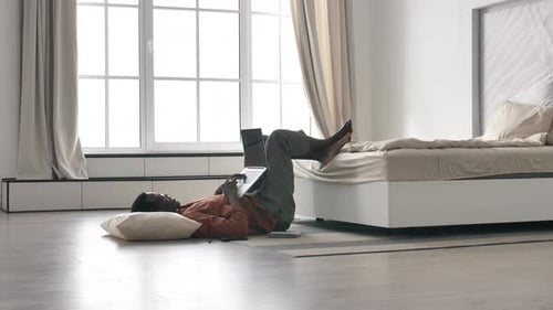 African American Freelancer Works at Home on a Laptop Lying on the Floor in Front of a Large Bright