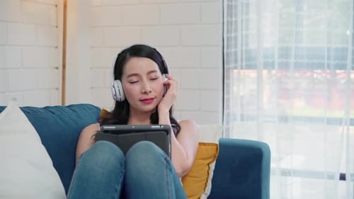 Asiatische Frau hört Musik und benutzt Tablet, Frau nutzt Entspannungszeit, wenn sie auf dem heimischen Sofa liegt.