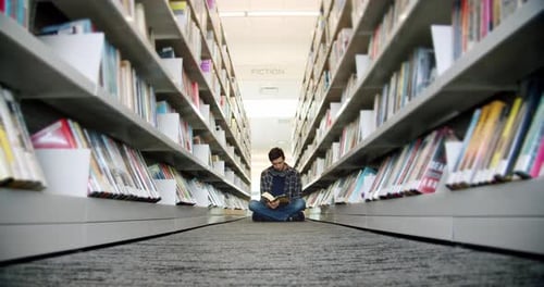 College Student Sitting on the Floor in the Library and Reading a Book Between Rows of Books