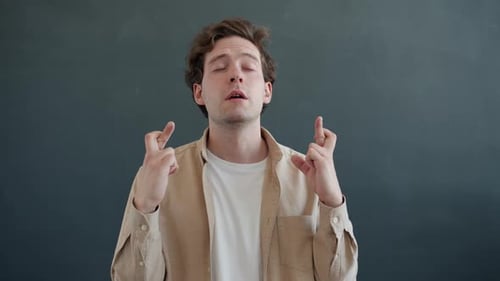 Portrait of Hopeful Man Crossing Fingers and Closing Eyes Praying Standing on Gray Color Background