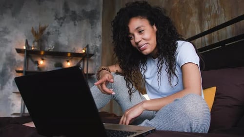 Junge attraktive afroamerikanische Frau nutzt Laptop für einen Videoanruf im Bett winkend