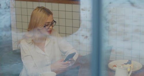 Attractive Young Businesswoman in Glasses Using Smartphone in Cafe View Through the Window