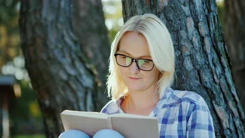 A Blonde Young Woman in Glasses Reads a Book in the Park