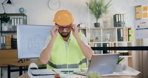 Architect in Helmet and Vest Sitting at the Table in Design Office and Working with Paper Plan