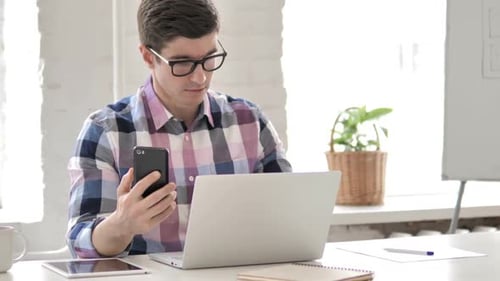 Young Adult Using Smartphone at Desk with Laptop