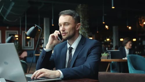 Cheerful Young Man in Formalwear Talking on Mobile Phone and Using Laptop in Cafe