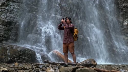 Un homme de voyage avec un sac à dos jaune debout sur le fond d'une cascade fait un paysage photo