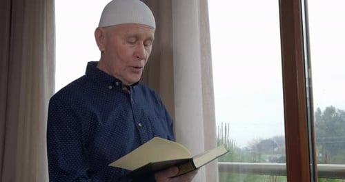 Senior Man Reads Religious Book Indoors by Window