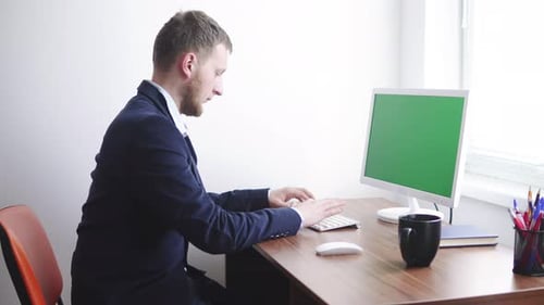 Young Man is Working on a Computer with a Mockup Green Screen at the Table By the Window