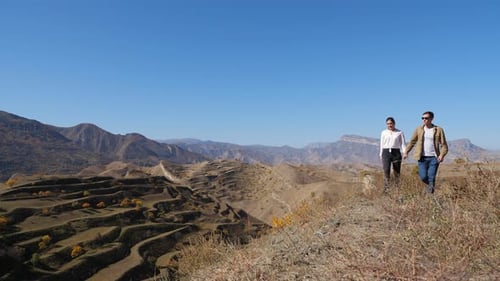 Couple Walks Along Hill with Dry Grass Against Cascade Slope
