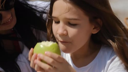 Mom and daughter eat an apple and laugh on Family picnic near sea on sunny summer day.