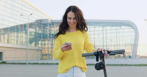 Mujer que usa su teléfono mientras camina con un scooter eléctrico cerca de un gran edificio urbano en verano