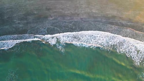 Aerial view of bluish green foamy waves breaking on sandy beach, while sun rays shines over the wate