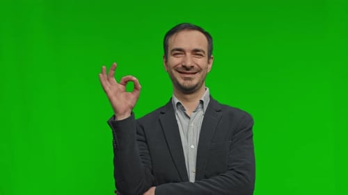 Young Handsome Man Making OK Sign Over Green Background and Smiles to Camera