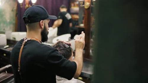 Side and Back View of the Stylish Barber with Beard Performing a Haircut with Scissors Holding a