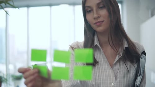 A Young Woman in a Business Suit Sticks Sticky Sheets with Reminders on a Glass Partition in the