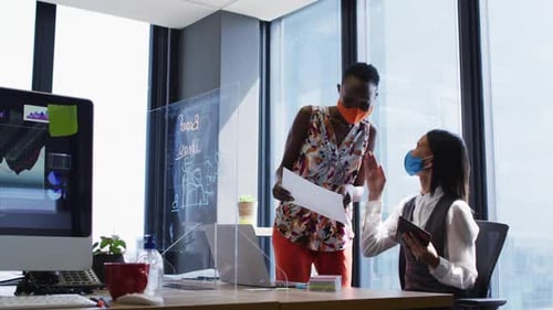Diverse female office colleagues wearing face masks discussing at modern office