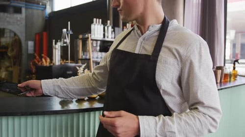 Portrait of caucasian man wearing apron working at a bar, using tablet and smiling to camera