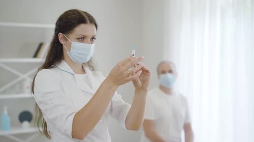 Serious Beautiful Woman in Face Mask Preparing Injection for Patient Sitting in Hospital Ward at the