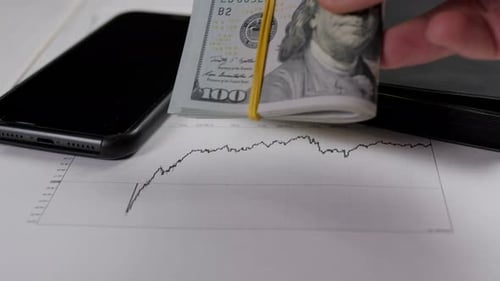 A Man Puts a Stack of Dollar Bills on a Table with a Phone and Financial Charts