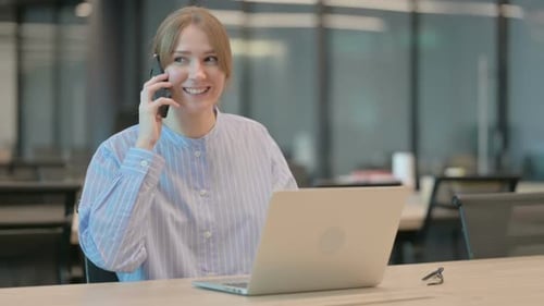 Young Woman Talking on Smartphone While Using Laptop in Office