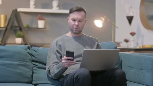 Man Using Phone and Laptop on Couch Indoors