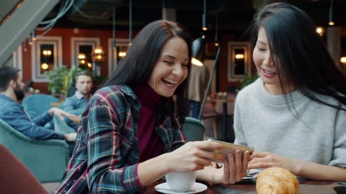 Joyful Female Friends Looking at Smartphone Screen in Cafe Talking Laughing Having Fun