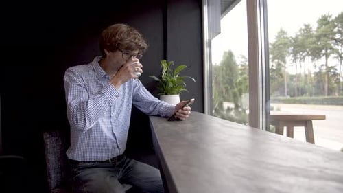 Man typing a message on the phone while in a cafe and drinking a coffee