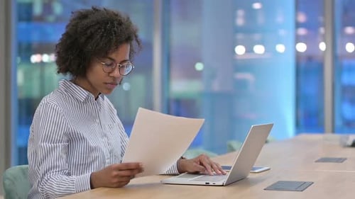 Focused African Businesswoman with Laptop Reading Documents in Office