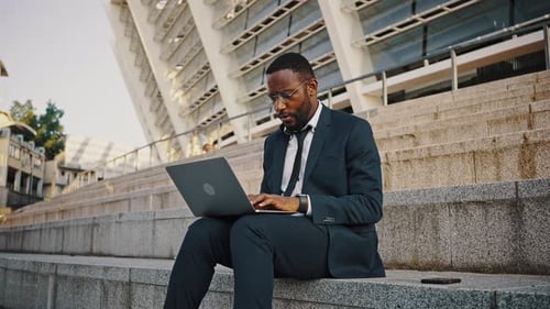 Focused African American Man Freelancer in Eyeglasses Typing on Laptop Watching on Smartphone and
