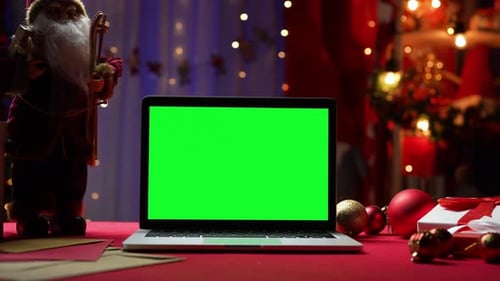 Laptop with Green Screen Chroma Key Stands on Red Table Next to Santa Claus Gifts and Toys
