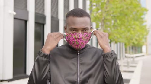 A Young Black Man Puts on a Face Mask and Looks at the Camera - an Office Building