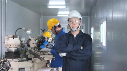 Factory worker with white hardhat and blue uniform stand with arm-crossed in front of co-worker
