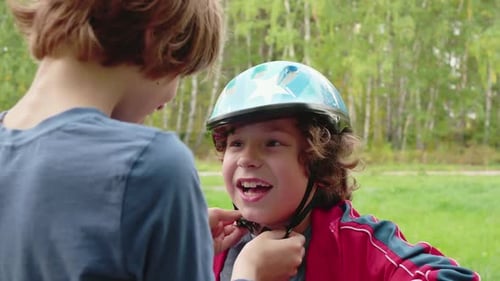 Older Brother Putting Bicycle Helmet on Little Brother Head in Summer Park