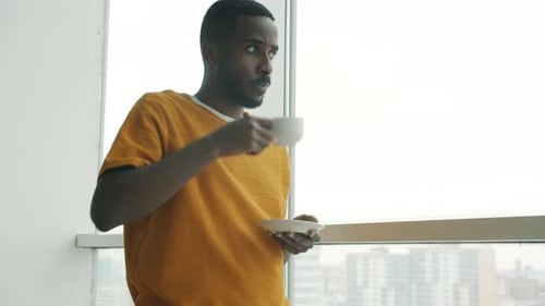 African American Man Drinking Coffee and Watching City From Panoramic Window Standing Alone in