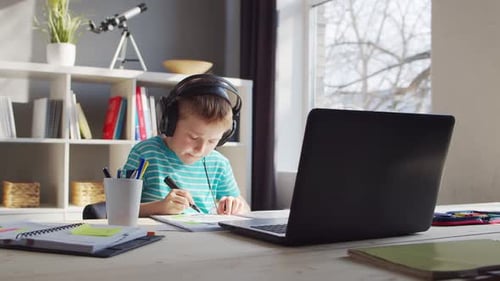 Boy with Headphones Writing in Notebook at Desk
