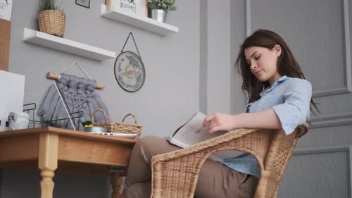Girl Designer Leafs Through a Magazine While Sitting at a Table in a Home Studio