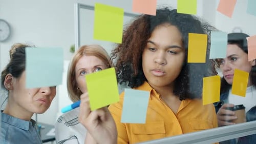 Group of Women Talking and Writing on Colorful Sticky Notes Working with Glassboard in Office