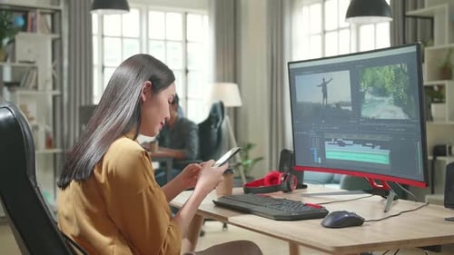 Asiatische Videoredakteurin arbeitet und benutzt Handy mit ihrem PC im Büro