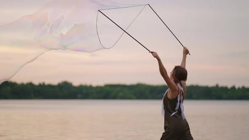 A Young Female Artist Shows a Soap Bubble Show Blowing Up Huge Soap Bubbles on the Shore of a Lake