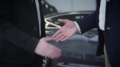 Close-up of Two Unrecognizable Caucasian Men Shaking Hands in Front of Automobiles in Car Dealership