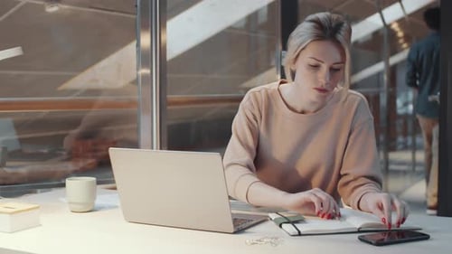 Blonde Female Office Worker Using Laptop and Taking Notes at Desk