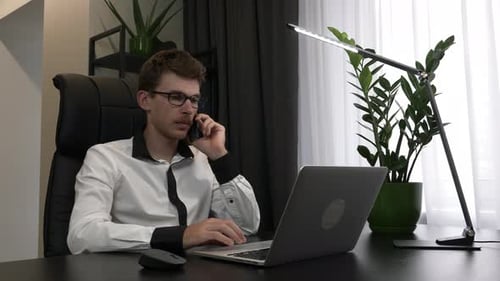 Focused man in eyeglasses is speaking by mobile phone in his modern business office.