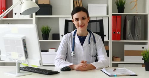Arzt im weißen Laborkittel sitzt am Arbeitsplatz im hellen Büro und schaut in die Kamera
