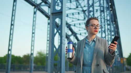Woman in a Coat Walking Around the City in the Early Morning Drinking Coffee and Using Smartphone